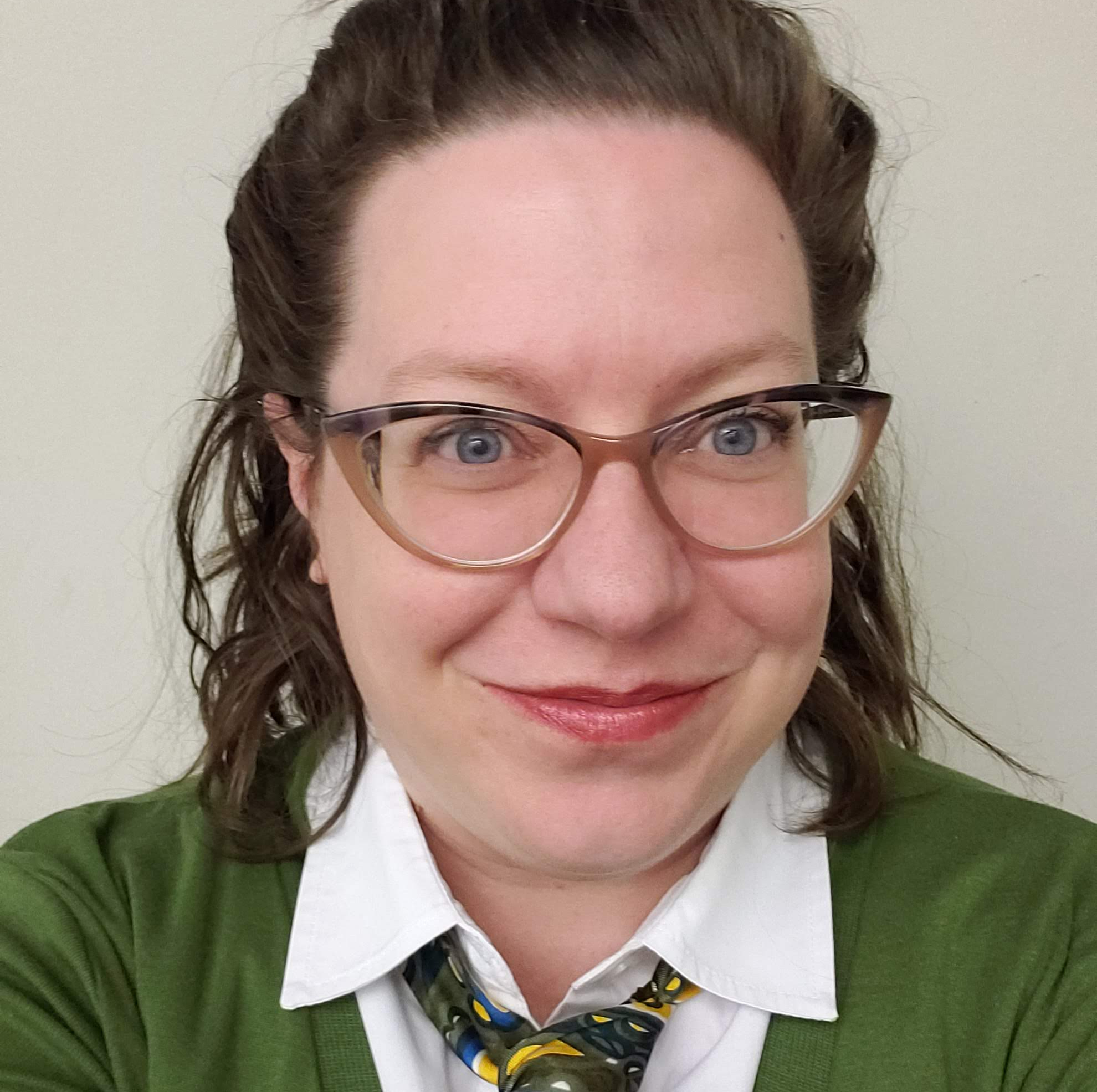 A smiling woman with glasses, wearing a green sweater over a white shirt with a multicolored tie.