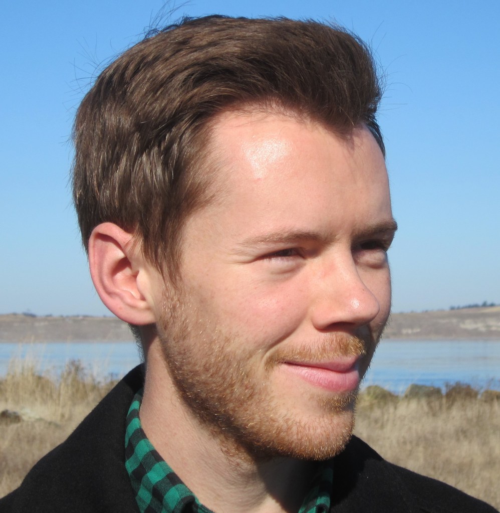 A smiling man looking into the distance. He has short brown hair, a short beard and mustache, and is wearing a green checked shirt with a black jacket. In the background, barren land and water can be seen.